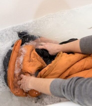 Person hand-washing an orange backpack in a sudsy bathtub