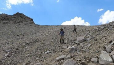 Two hikers descend a steep, loose scree slope beneath a blue sky