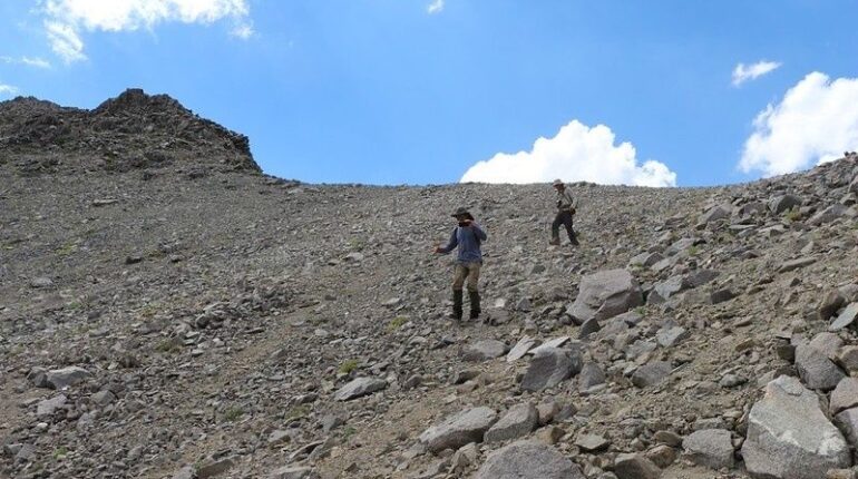 Two hikers descend a steep, loose scree slope beneath a blue sky