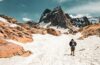 Hiker crossing a snowy mountain trail beneath a steep rocky peak