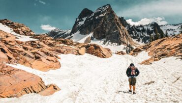 Hiker crossing a snowy mountain trail beneath a steep rocky peak